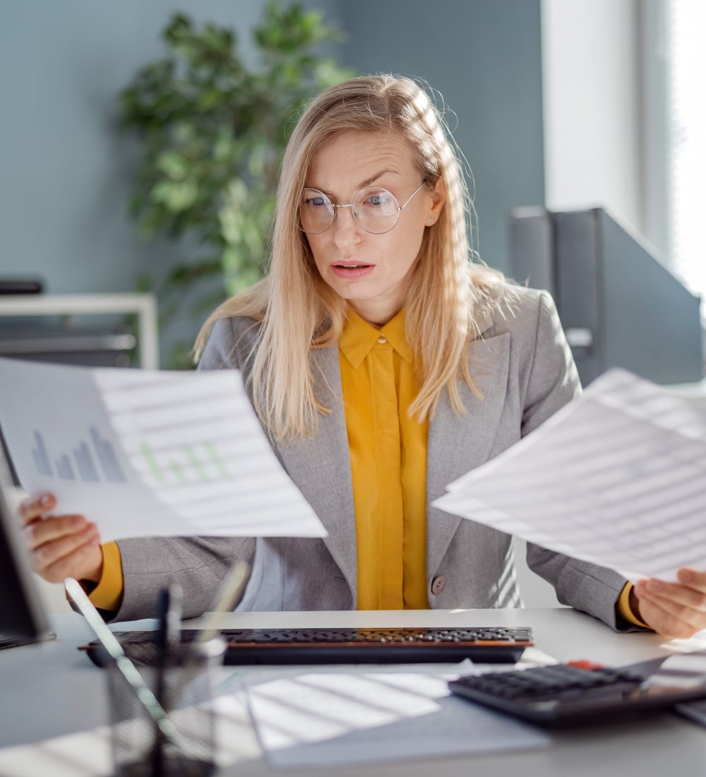 Mature caucasian lady in eyeglasses and formal suit examining various documents while sitting at desk with modern pc. Concept of people, work and gadgets.